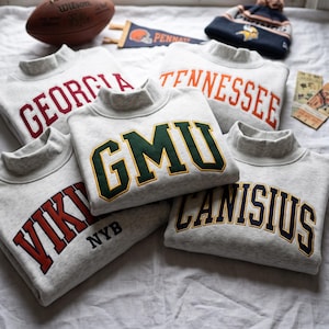 Könnte beinhalten: Fünf hellgraue Sweatshirts mit Stehkragen und College-Schriftzügen in verschiedenen Farben, darunter Georgia, Tennessee, GMU, Vikings NYB und Canisius. Ein brauner Wilson-Football, ein Wimpel und eine Strickmütze sind ebenfalls zu sehen.