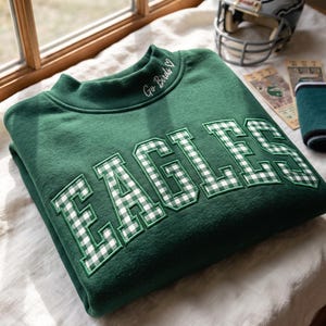 May include: A dark green crewneck sweatshirt featuring the word EAGLES in white and green gingham block letters across the chest. The collar includes embroidered text that reads Go Birds with a small heart symbol above the lettering.