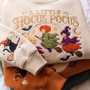 Może przedstawiać: Kremowa bluza z wyhaftowanym napisem A LITTLE HOCUS POCUS nad kolistą sceną przedstawiającą trzy czarownice na miotłach. Na rękawie znajduje się czarny kot na miotle. Poniżej znajdują się dwie dodatkowe bluzy w kolorze pomarańczowym i zielonym.
