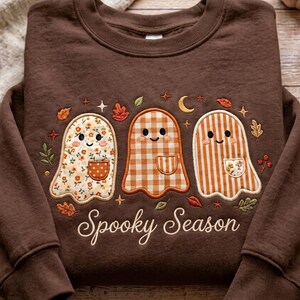 Op de afbeelding: Een donkerbruine sweater met ronde hals en een borduursel van drie spoken met kleine zakjes. De spoken hebben bloemen-, ruit- en streepmotieven. De tekst Spooky Season is eronder geborduurd in een wit cursief lettertype.