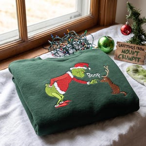 Könnte beinhalten: Ein dunkelgrüner Rundhals-Pullover mit einer Stickerei, die eine grüne Figur im roten Weihnachtsmannkostüm zeigt, die die Nase eines braunen Rentiers berührt. Die Stickerei enthält das Wort Boop in weißem Garn neben dem Finger der Figur.