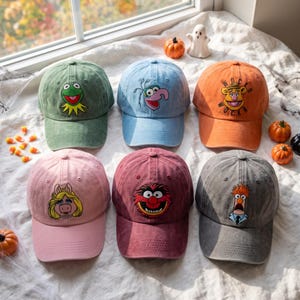 May include: Six baseball caps in green, light blue, orange, pink, maroon, and grey, each featuring an embroidered Muppet character. The green cap shows Kermit, light blue shows Gonzo, orange shows Fozzie Bear with Wocka Wocka text, pink shows Miss Piggy, maroon shows Animal, and grey shows Beaker.