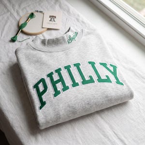May include: A light grey heathered sweatshirt featuring the word PHILLY in large, dark green embroidered block letters across the chest. The name Sophia is embroidered in green cursive on the mock neck collar. A wooden embroidery hoop with green thread and a small card with a bell icon are visible nearby.
