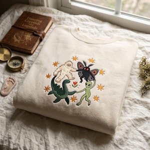 Peut inclure: Un sweat-shirt à col rond couleur crème avec une broderie représentant un yéti blanc, une créature ailée noire, un monstre marin vert et un extraterrestre vert se tenant la main autour d'une petite fleur rouge, entourés d'étoiles jaunes.