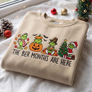 Peut inclure: Un sweat-shirt à col rond beige avec une broderie représentant quatre personnages verts dans des tenues saisonnières. Le texte indique THE BER MONTHS ARE HERE. Le motif inclut des feuilles d'automne, une citrouille, un chapeau et un sapin.