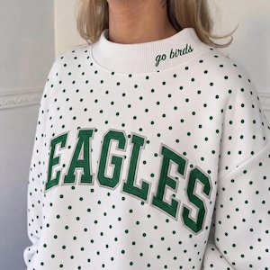Op de afbeelding: Een wit sweatshirt met een groen stippenpatroon en een geribbelde opstaande kraag. Het woord EAGLES is in grote groene blokletters op de borst geborduurd en de tekst go birds is in het groen op de kraag geborduurd.