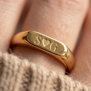 May include: A gold signet ring featuring a smooth, polished surface with an engraved design consisting of the letter S, a small heart symbol, and the letter G centered on the flat top portion of the band, creating a clear and legible inscription.