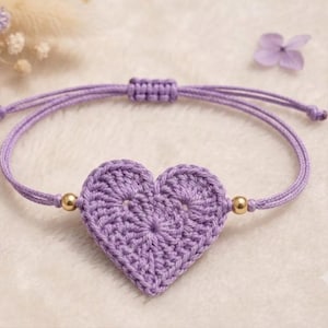 Könnte beinhalten: Ein Armband aus dünner violetter Kordel mit einem lavendelfarbenen Häkelherz als Anhänger. Das Armband ist mit zwei kleinen goldfarbenen Metallperlen neben dem Herz und einem verstellbaren Schiebeknoten aus der gleichen Kordel versehen.
