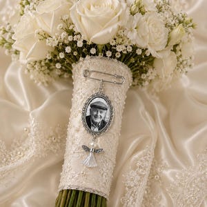 Pode incluir: Um buquê de noiva de rosas brancas e gipsófila com o cabo envolto em renda. Um alfinete de segurança prateado prende um pingente oval com a foto de uma pessoa idosa de boina, acentuado por um pequeno pingente de anjo branco com pérola.