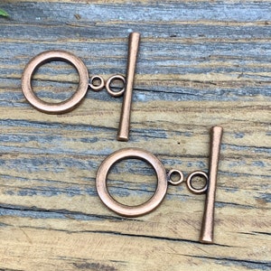 May include: Two copper-toned metal toggle clasps with a round ring and a straight bar. The clasps are on a wooden surface.