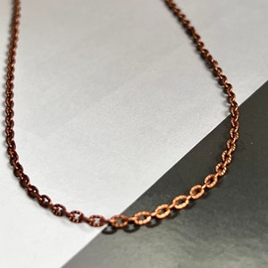 May include: A copper chain necklace with a delicate, oval-shaped link design. The chain is lying on a white surface.