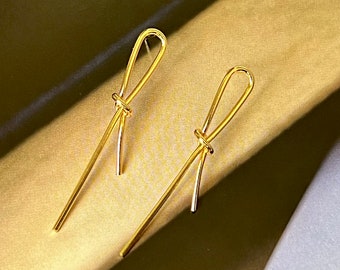 Könnte beinhalten: Ein Paar goldene Knotenohrringe. Die Ohrringe sind lang und dünn, mit einem einfachen Knotendesign.