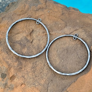 May include: Two silver-toned hoop earrings with a hammered texture. Each earring has a small loop at the top for attachment. The earrings are circular and have a rustic, handmade appearance. The background is a natural stone surface.