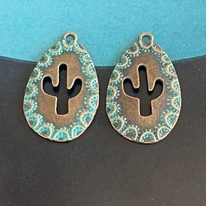 May include: Two copper-colored teardrop-shaped earrings. Each earring features a cutout cactus design in the center. The edges are adorned with a turquoise-colored, patterned border. The earrings have a small loop at the top for hanging.