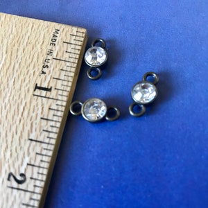 May include: Three antique bronze-colored metal connector charms with clear round rhinestones. The charms are approximately 1/2 inch in diameter and are shown on a wooden ruler.