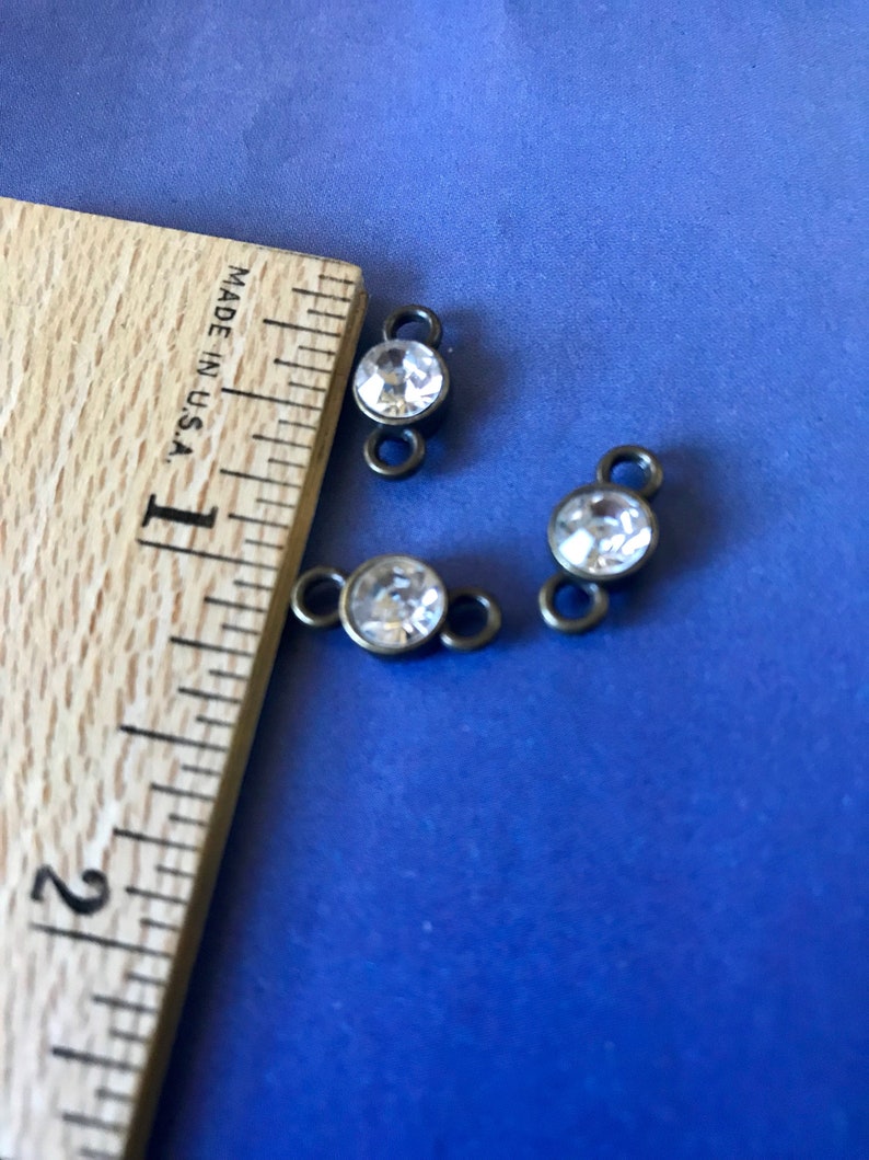 May include: Three antique bronze-colored metal connector charms with clear round rhinestones. The charms are approximately 1/2 inch in diameter and are shown on a wooden ruler.