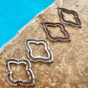 May include: Four metal pendant charms in the shape of a quatrefoil. The charms are in three different finishes: silver, copper, and a darker copper. The charms are arranged in a row on a brown, textured surface.