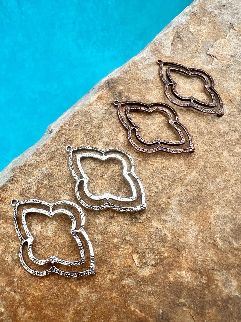 May include: Four metal pendant charms in the shape of a quatrefoil. The charms are in three different finishes: silver, copper, and a darker copper. The charms are arranged in a row on a brown, textured surface.