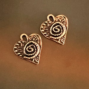 May include: Two copper heart-shaped charms with a swirl design. Each charm has a loop at the top for attaching to jewelry or other crafts.