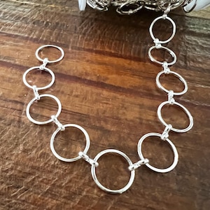 May include: A silver-colored chain necklace with large, open circular links. The necklace is displayed on a wooden surface, with a roll of chain visible in the background. The chain is made of metal and has a shiny finish.