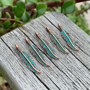 May include: Six copper-colored feather charms with turquoise enamel. The feathers are arranged in a row on a wooden surface.