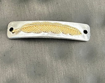 Puede incluir: Una barra de metal plateado con un diseño de pluma dorada. La barra tiene dos agujeros en cada extremo.