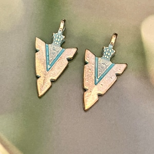 May include: Two gold-colored arrowhead pendants with a turquoise and white enamel design. The arrowheads have a pointed tip and a loop at the top for attaching to a chain or cord.