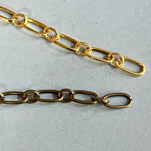 May include: Close-up of two gold-colored chain necklaces. The top chain has a bright, shiny finish, while the bottom chain has a darker, antique finish. Both chains feature oval links.