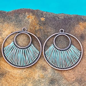 May include: A pair of copper-colored hoop earrings with a circular design. Each earring features a smaller inner circle and a fan-shaped arrangement of light blue and silver threads. The earrings are displayed on a textured, brown surface.
