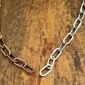 May include: Close-up of two sections of a chain necklace. One section is a copper color, and the other is silver. The chain links are oval-shaped and lie on a wooden surface.