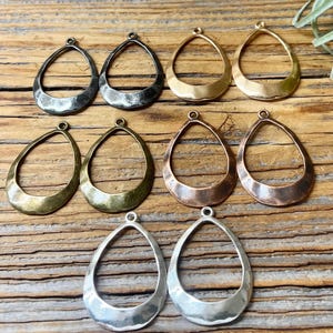 May include: A collection of ten teardrop-shaped metal jewelry charms arranged in pairs. The charms feature a smooth, open-center design with a hammered texture at the base. The set includes various metallic finishes, including silver, gold, copper, bronze, and dark gunmetal tones, each with a small loop at the top for attachment.