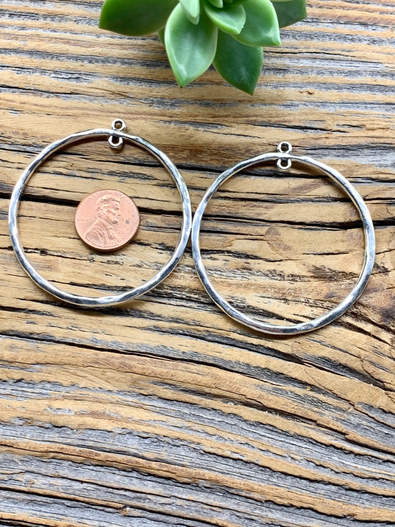 Puede incluir: Dos pendientes de aro de plata con acabado martillado. Los aros son grandes y tienen un di&aacute;metro de unos 5 cm. Los pendientes se muestran sobre una superficie de madera.