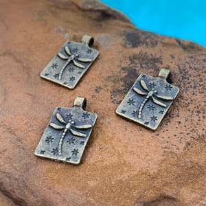 May include: Three antique gold-toned metal charms shaped like rectangles with a dragonfly design and a star pattern.