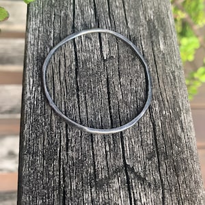 May include: A simple silver bangle bracelet, made of a thin, round wire, is shown on a wooden surface.