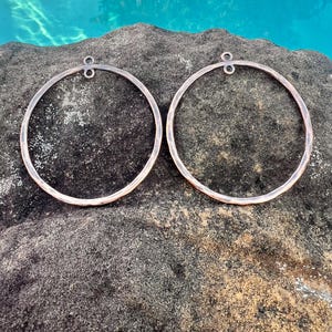 May include: Two large, hammered copper hoops with a simple loop at the top. The hoops are lying on a dark gray rock surface.