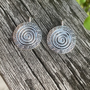 May include: Two circular silver-toned metal pendants featuring a central spiral design surrounded by a geometric Greek key border. Each pendant has a small metal loop at the top for attachment, with a dark oxidized finish in the recessed areas.