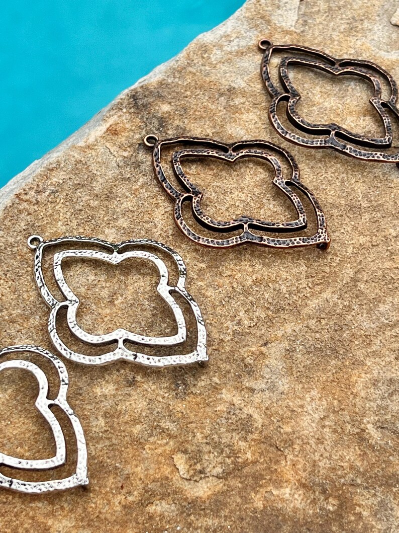 May include: Four metal pendant charms in different finishes: copper, antique copper, silver, and antique silver. Each charm features a double-layered quatrefoil design with a hammered texture.