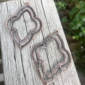 May include: Two antique copper-colored metal pendant charms shaped like a stylized flower with a double-layered design.