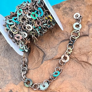 May include: A spool of colorful chain made from metal rings and washers. The chain is a mix of copper, silver, and turquoise colors. The chain is on a white spool and is laying on a brown rock.