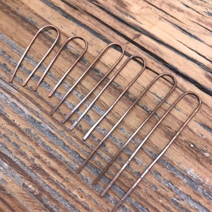 May include: Seven copper hair pins with a simple, curved design. The pins are arranged in a row on a wooden surface.
