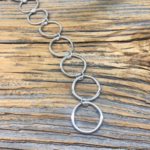 May include: A silver-colored chain necklace featuring a series of interconnected, open circles. The necklace is displayed against a rustic, weathered wooden background, with a hint of green foliage in the upper right corner.