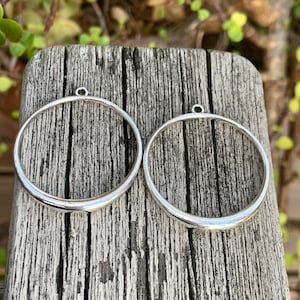 K&ouml;nnte beinhalten: Zwei silberfarbene Metallreifen mit einer kleinen Schlaufe oben. Die Reifen liegen auf einer Holzoberfl&auml;che.