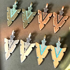 May include: Eight metal arrowhead pendants arranged in two rows. The top row features two weathered green-toned pendants followed by two copper-toned pendants. The bottom row includes two silver-toned pendants and two gold-toned pendants with blue accents.