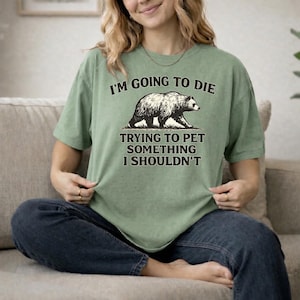 May include: A sage green short-sleeve t-shirt featuring a black graphic of a bear. The text above the bear reads I'M GOING TO DIE and the text below the bear reads TRYING TO PET SOMETHING I SHOULDN'T. The shirt has a crew neckline and a relaxed fit.