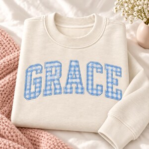 May include: A cream-colored crewneck sweatshirt featuring the word GRACE in large, block-style letters. The letters are filled with a light blue and white gingham check pattern and are outlined with a stitched border across the chest.