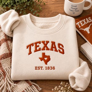 May include: A cream-colored crewneck sweatshirt featuring the word TEXAS in arched, burnt orange lettering. Below the text is a burnt orange silhouette of the state of Texas with a heart cutout, followed by the text EST. 1836 in matching orange.