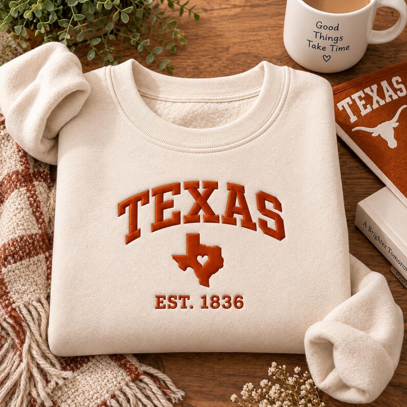 May include: A cream-colored crewneck sweatshirt featuring the word TEXAS in arched, burnt orange lettering. Below the text is a burnt orange silhouette of the state of Texas with a heart cutout, followed by the text EST. 1836 in matching orange.