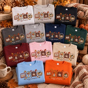 May include: A collection of crewneck sweatshirts in various colors, each featuring a graphic print of four framed autumn-themed illustrations including a book, a vase of leaves, a coffee mug, and a house, with the text cozy season below.