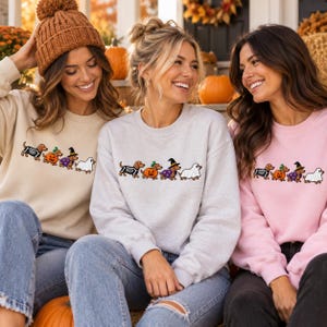 May include: Three crewneck sweatshirts in beige, light gray, and light pink, each featuring an embroidered graphic of four dachshunds in Halloween costumes: a skeleton, a pumpkin, a witch, and a ghost, arranged in a horizontal line across the chest.