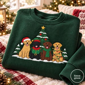 May include: A forest green crewneck sweatshirt featuring an embroidered design of four dogs sitting in front of a Christmas tree. The dogs include a golden retriever in a Santa hat, a black dog in an elf costume, a brown dog with a green scarf, and a golden retriever wrapped in colorful string lights.